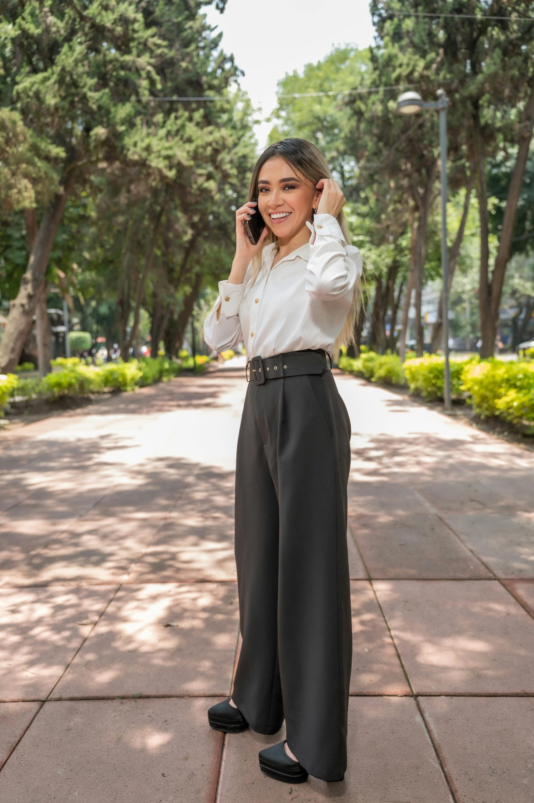 Elegant woman in business attire talking on the phone in a serene park setting.